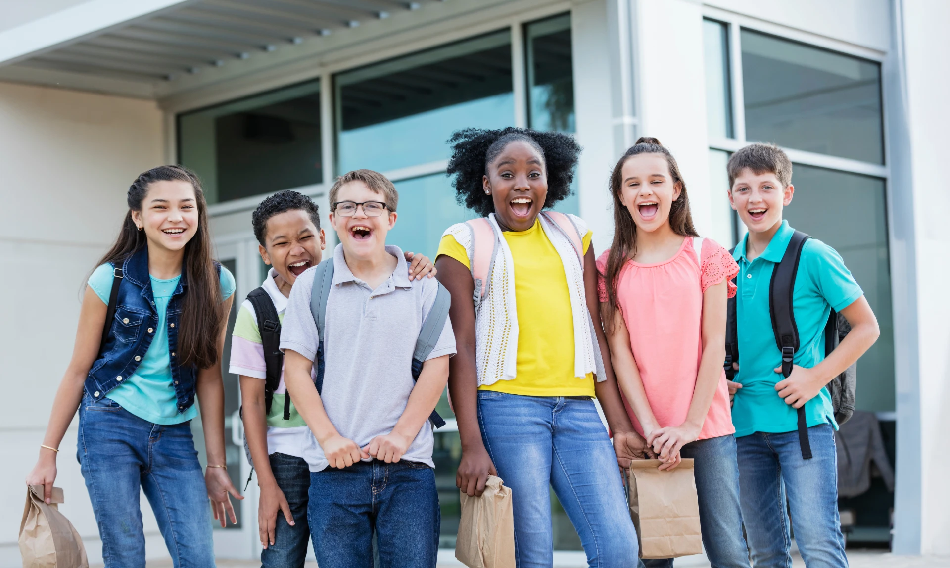 Six diverse children smiling and laughing outside a school building, some carrying backpacks and brown paper lunch bags.